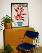Poster moderno con pianta stilizzata rossa con occhi in vaso blu, stampa artistica contemporanea con scritta “Cry for something that can grow”, decorazione per interni con mobili in legno e piante verdi