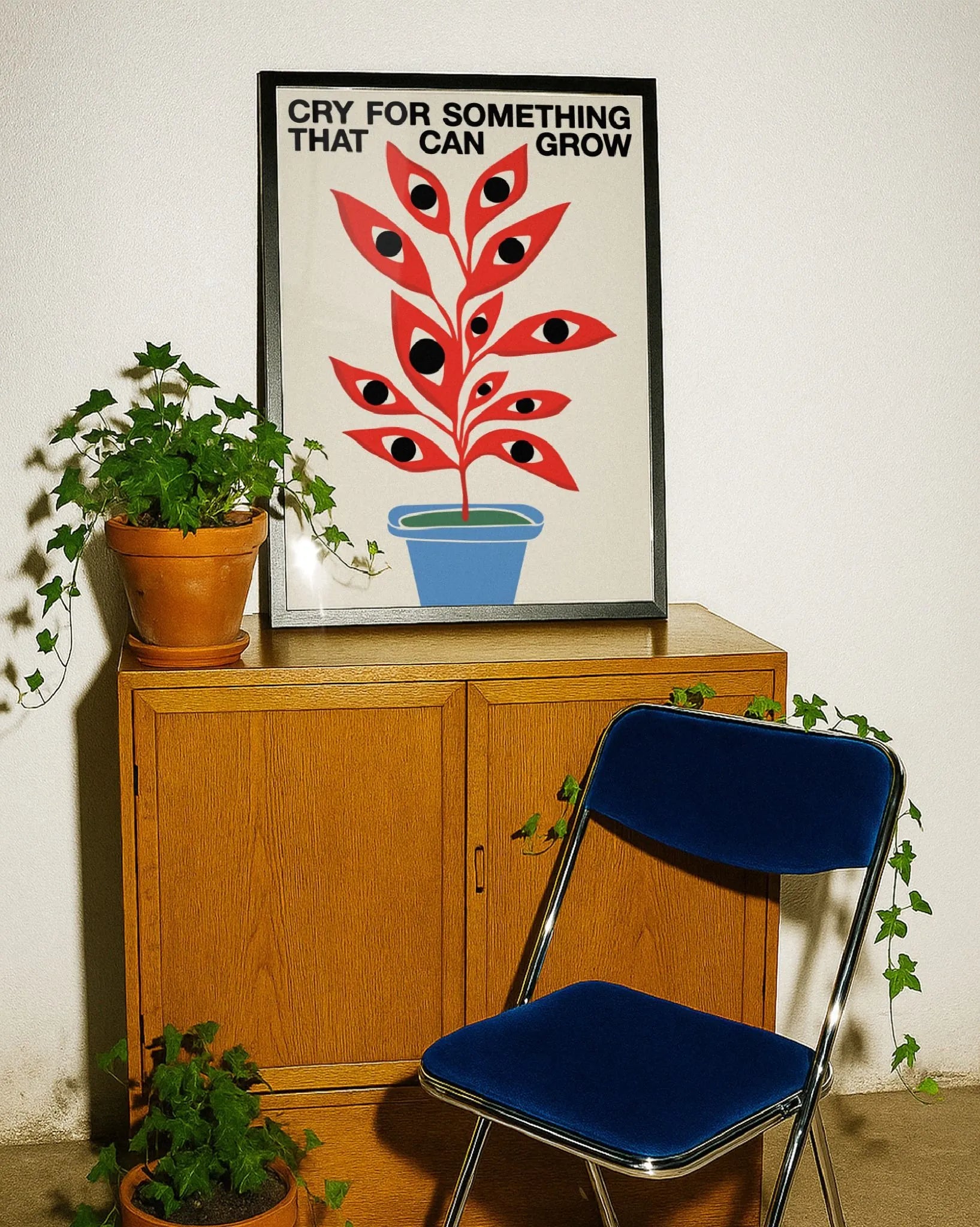 Poster moderno con pianta stilizzata rossa con occhi in vaso blu, stampa artistica contemporanea con scritta “Cry for something that can grow”, decorazione per interni con mobili in legno e piante verdi